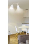 Lampa sufitowa Atlas 2-punktowa w kolorze białym, z dwoma kloszami w kształcie stożka, zamontowana na prostokątnej podstawie. Ujęcie z góry, w tle nowoczesna kuchnia z białymi meblami i okrągłym stołem.