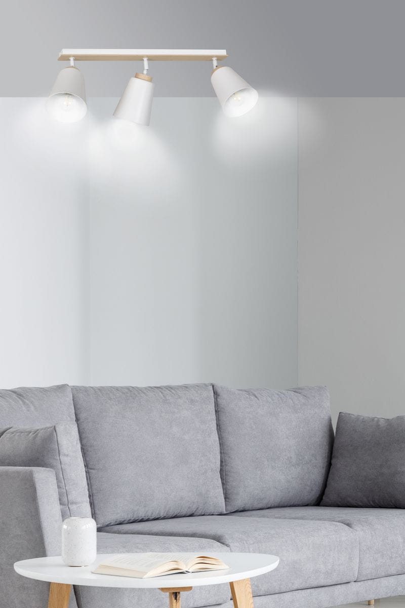 Lampa sufitowa Atlas 3-punktowa w kolorze białym z drewnianymi detalami. Ułożona poziomo, z trzema kloszami skierowanymi w dół. Tło w odcieniach szarości, w aranżacji z sofą i stolikiem. Oświetlenie włączone, emitujące jasne światło.
