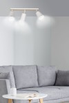 Lampa sufitowa Atlas 3-punktowa w kolorze białym z drewnianymi detalami. Ułożona poziomo, z trzema kloszami skierowanymi w dół. Tło w odcieniach szarości, w aranżacji z sofą i stolikiem. Oświetlenie włączone, emitujące jasne światło.