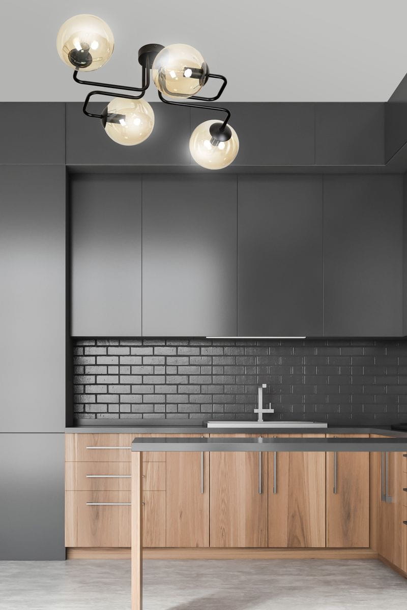 Brendi lampa sufitowa 4-punktowa w kolorze miodowym. Cztery szklane klosze w kształcie kul, zamocowane na czarnym stelażu w kształcie falistym. Lampa wisząca zainstalowana w nowoczesnej kuchni z ciemnymi szafkami i drewnianymi elementami.