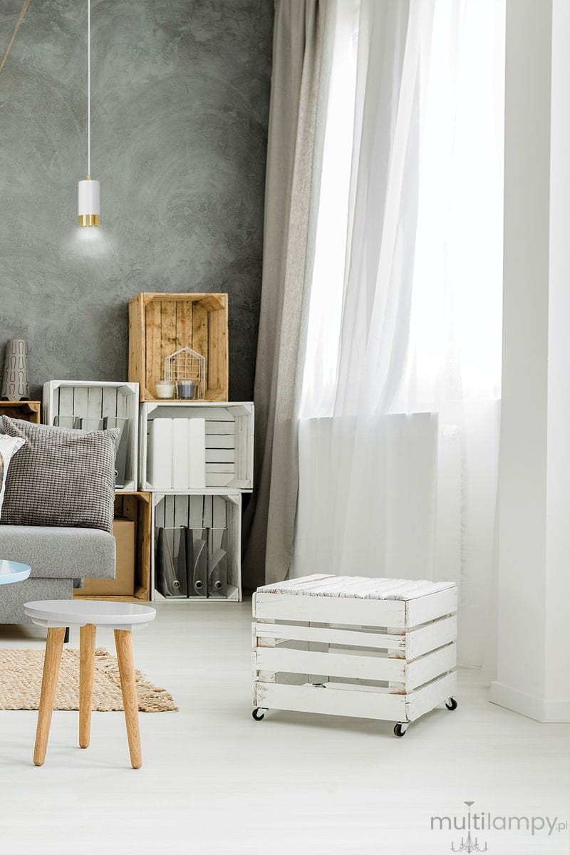 Lampa wisząca Fumiko 1-punktowa w kolorze białym z złotymi detalami, zawieszona na cienkim kablu. Widoczna w jasnym wnętrzu z szaro-białymi ścianami, obok mebli w stylu skandynawskim oraz drewnianymi skrzynkami.