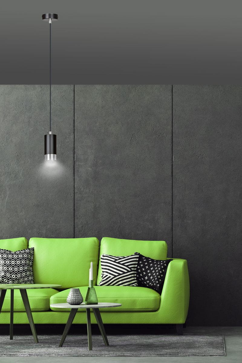 Lampa wisząca Fumiko 1-punktowa w kolorze czarnym z chromowanym detalem, zawieszona na cienkim przewodzie, o cylindrycznym kształcie. W tle widoczna zielona sofa z poduszkami w geometryczne wzory, na szarym tle.