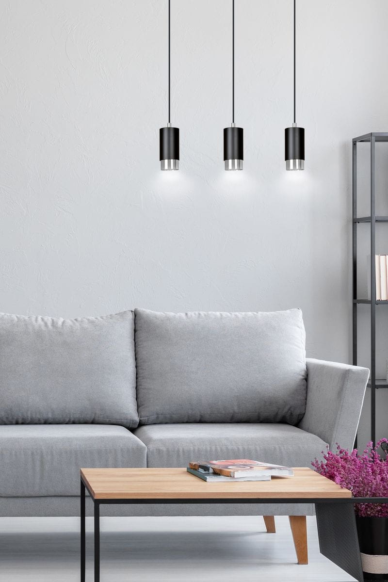 Lampa wisząca Fumiko 3-punktowa w kolorze czarnym i chromowanym, z trzema cylindrycznymi kloszami. Zawieszona na cienkich linkach, w nowoczesnym wnętrzu z szarym sofą i drewnianym stolikiem. Tło jasne, minimalistyczne.