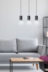 Lampa wisząca Fumiko 3-punktowa w kolorze czarnym i chromowanym, z trzema cylindrycznymi kloszami. Zawieszona na cienkich linkach, w nowoczesnym wnętrzu z szarym sofą i drewnianym stolikiem. Tło jasne, minimalistyczne.