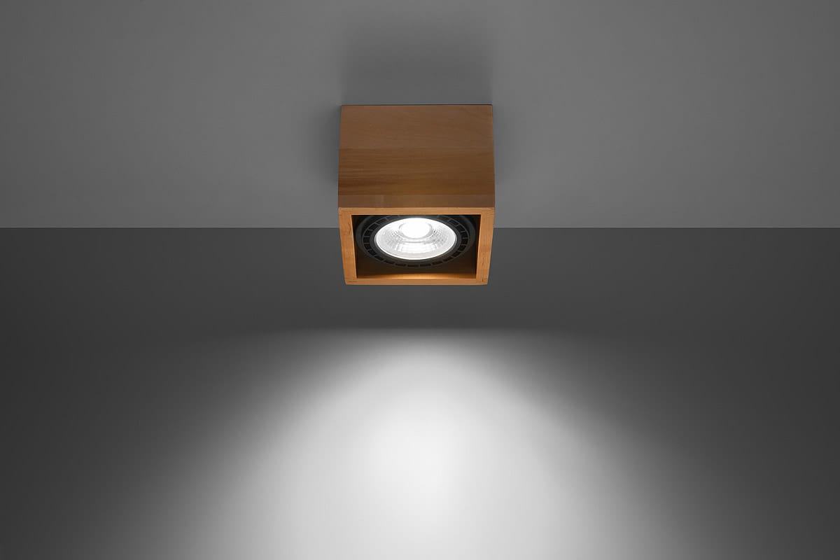Lampa sufitowa Quatro 1-punktowa w kolorze drewna, o kwadratowej formie. Zainstalowana na suficie, z widocznym reflektorem wewnętrznym. Oświetlenie rzuca jasne światło na szare tło.