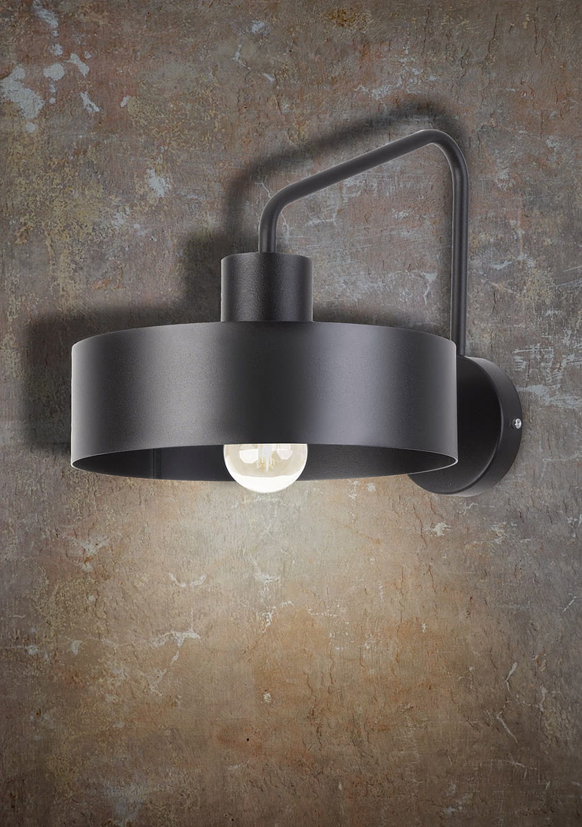 Czarny kinkiet jedno-punktowy Jumbo z metalowym kloszem w kształcie cylindrycznym, zawieszony na ścianie. Widoczna żarówka w środku, minimalistyczny design. Kąt ujęcia z boku, na tle teksturowanej, brązowej ściany.