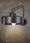 Czarny kinkiet jedno-punktowy Jumbo z metalowym kloszem w kształcie cylindrycznym, zawieszony na ścianie. Widoczna żarówka w środku, minimalistyczny design. Kąt ujęcia z boku, na tle teksturowanej, brązowej ściany.