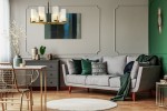 Lampa wisząca Fortano MOD089PL-08BS z ośmioma punktami świetlnymi, w metalowej, złotej oprawie z białymi kloszami. Ujęcie frontalne, lampa zawieszona nad szarym sofą w nowoczesnym wnętrzu. W tle zielona ściana i dekoracje.