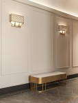 Kinkiet Verno Parette Old Gold w kolorze złotym, o ażurowym kloszu z geometrycznym wzorem, zamontowany na ścianie. Widoczny w minimalistycznym wnętrzu z elegancką ławką. Oświetlenie dwupunktowe tworzy przytulny klimat.