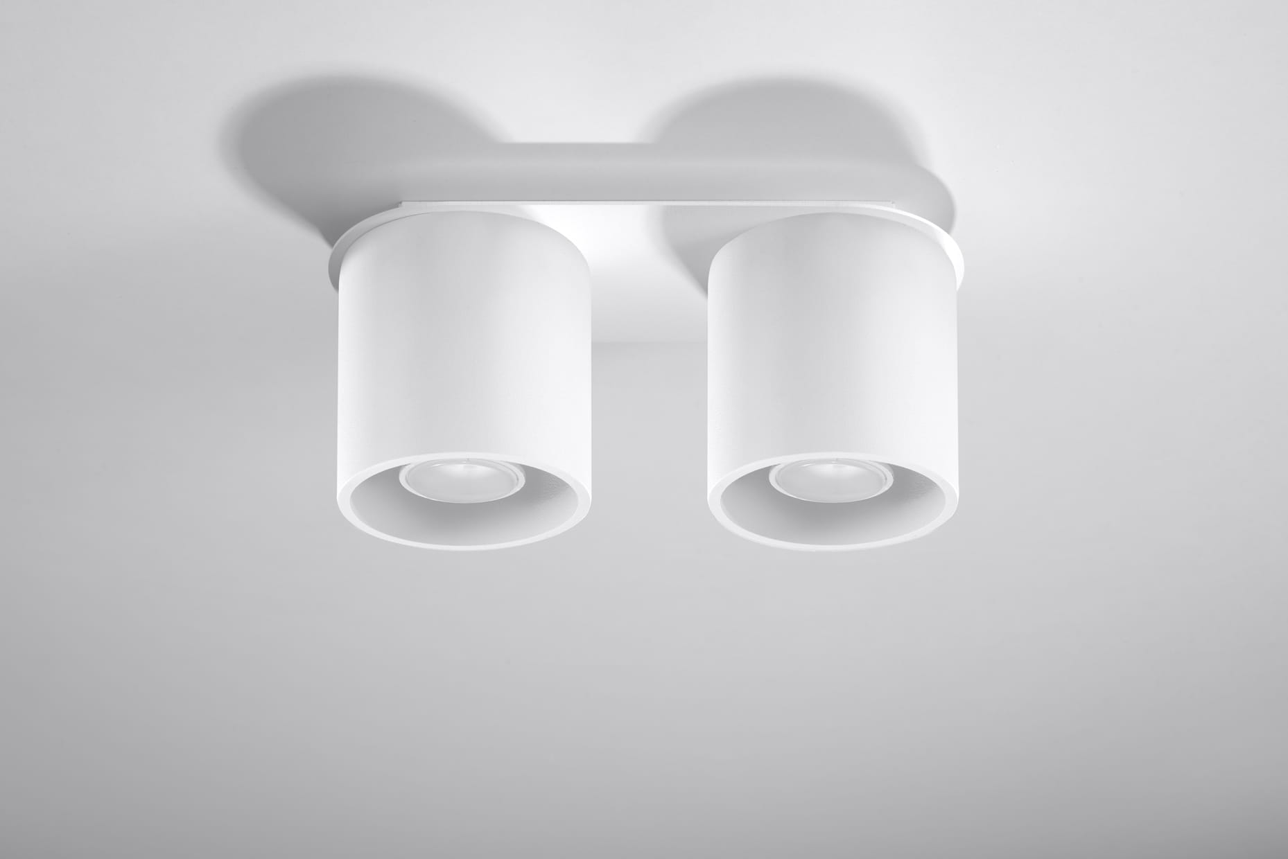 Biała lampa sufitowa Orbis z dwoma cylindrycznymi punktami świetlnymi. Produkt zamontowany na prostokątnej podstawie, widoczny z dołu. Gładka, matowa powierzchnia oraz minimalistyczny design. Idealna do nowoczesnych wnętrz.