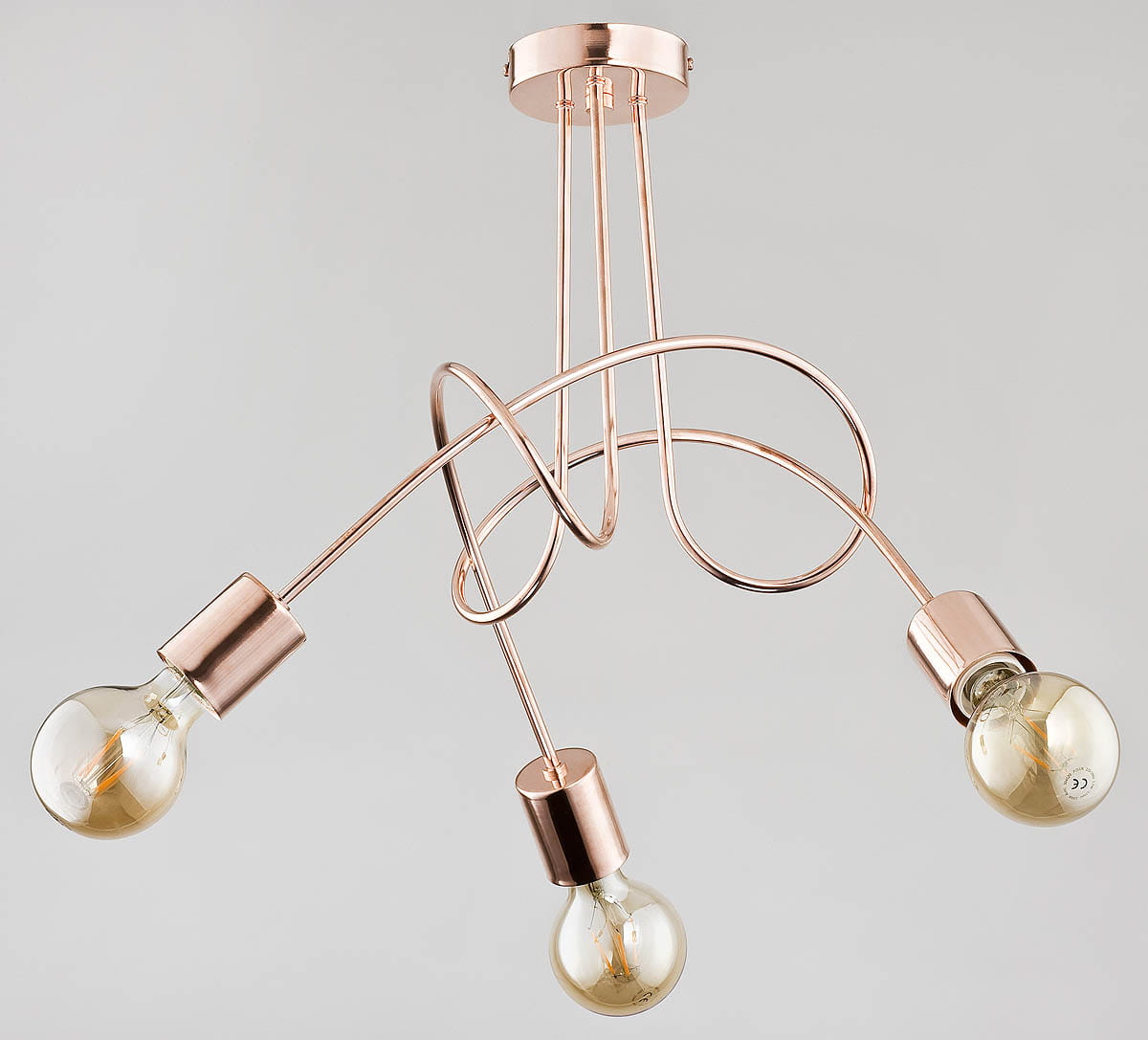 Lampa wisząca Tango 3-punktowa w kolorze miedzi z trójwymiarową konstrukcją. Posiada trzy źródła światła w stylowych oprawach, zawieszone na dekoracyjnych, zakrzywionych przewodach. Montaż sufitowy, na jednolitym tle.
