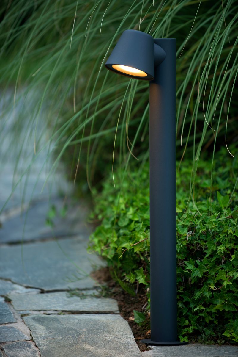 Lampa ogrodowa stojąca Dingo H70 w antracytowym kolorze, o prostym cylindrycznym kształcie. Ujęta pod kątem, z widocznym kloszem skierowanym w dół, zamontowana na podłożu w otoczeniu zieleni i kamiennej alejki.