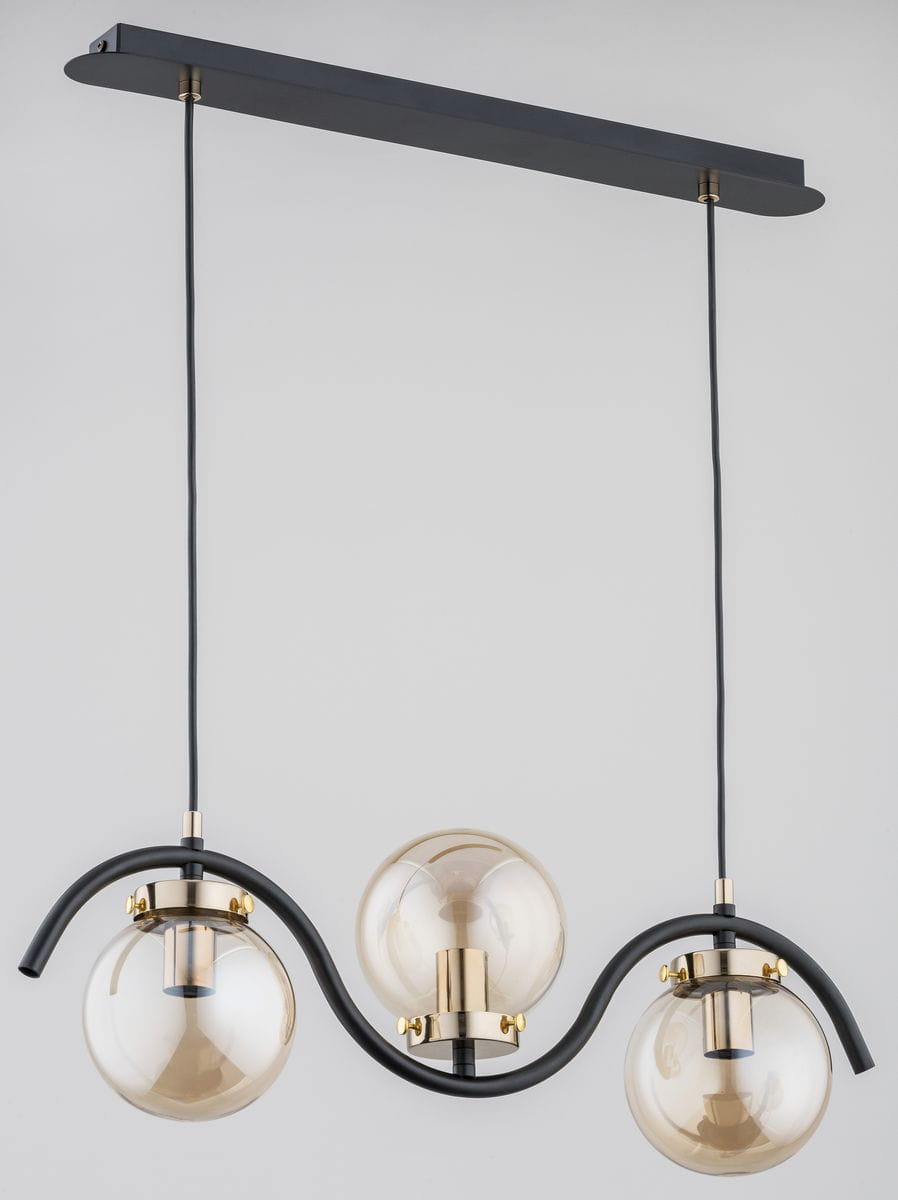 Lampa wisząca Korda 3-punktowa w złotym wykończeniu. Posiada trzy przezroczyste klosze w kształcie kul, zawieszone na czarnych przewodach. Metalowy uchwyt w kształcie fali, montowany do sufitu. Całość na jasnym tle.