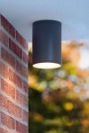 Lampa sufitowa elewacyjna Tubix 1-punktowa w kolorze antracytowym, cylindryczna, zawieszona pod sufitem, skierowana w dół. W tle widoczna ceglana ściana oraz rozmyte, kolorowe tło, co nadaje przyjemny klimat.