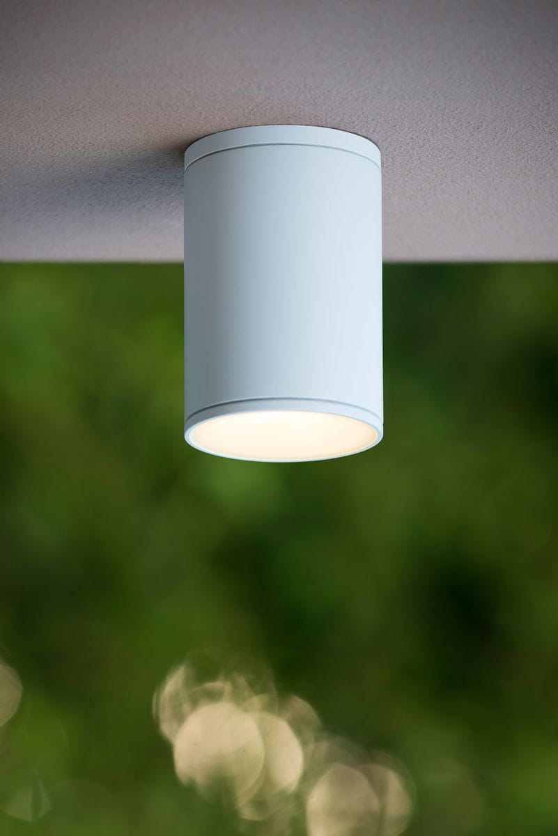 Biała lampa sufitowa elewacyjna Tubix, w kształcie cylindrycznym, montowana na suficie. Zbliżenie na górną część lampy z delikatnym źródłem światła, w tle rozmyte zieleń, co podkreśla jej zastosowanie na zewnątrz.