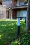 Lampa stojąca ogrodowa Dropa LED H59 z obudową w kolorze czarnym, z przezroczystą górą emitującą niebieskie światło. Umieszczona na trawniku, w tle nowoczesny budynek. Sterowana telefonem przez Bluetooth.