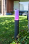Lampa stojąca ogrodowa Dropa LED H59 z czarną podstawą i przezroczystym kloszem, emitująca kolorowe światło, w tym fioletowe. Ujęcie z boku, umieszczona na trawie, w tle drewniana elewacja budynku.