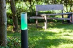 Lampa stojąca ogrodowa Dropa LED H59 w kształcie cylindrycznym, z czarnym podstawą i przezroczystą górą, emitująca zielone światło. Ustawiona na trawie, w tle drewniana ławka, otoczenie zielone. Sterowana telefonem przez Bluetooth.