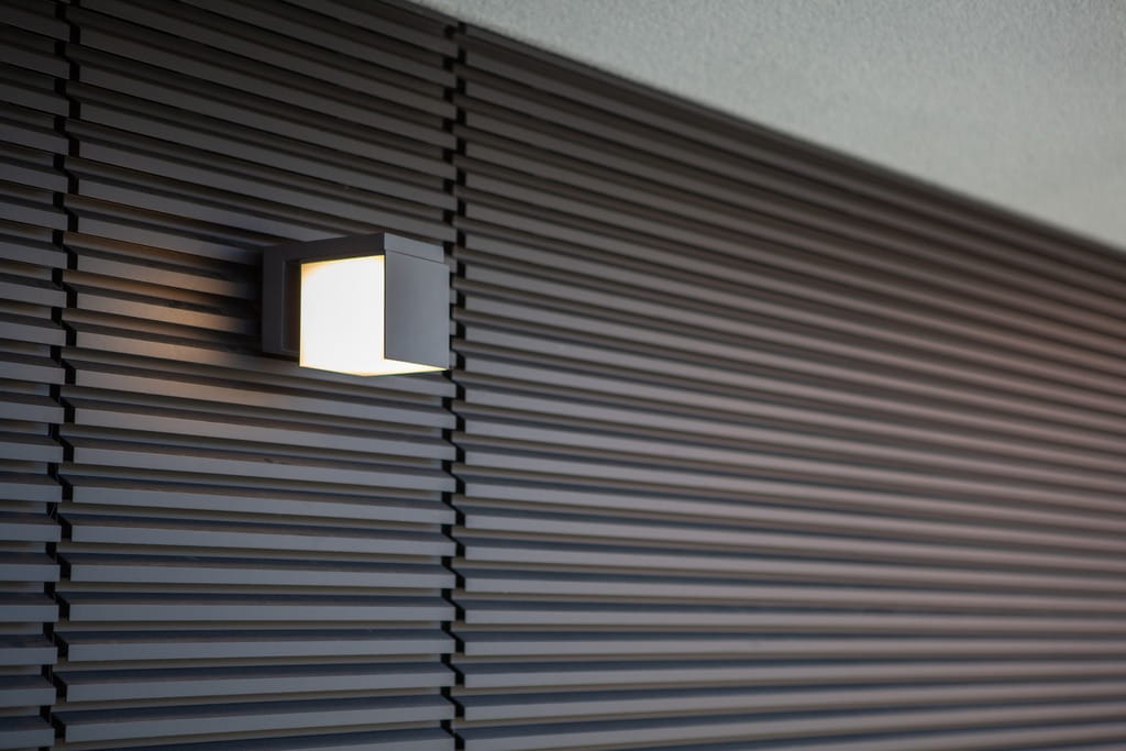 Cuba LED kinkiet elewacyjny 1-punktowy w kolorze czarnym, zamontowany na ścianie z poziomą fakturą. Kinkiet o kwadratowym kształcie, emitujący ciepłe światło, z regulowanym kątem oświetlenia. Tło w odcieniach szarości.