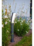 Lampa stojąca Kira LED H74, ciemnoszara, umieszczona w ogrodzie wśród kwiatów. Posiada prostokątny, zaokrąglony klosz w białym kolorze, stojącą podstawę i jest w pozycji pionowej. Tło z zielenią i kwiatami podkreśla jej elegancki design.