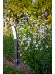 Lampa stojąca Kira LED H74 w ciemnoszarym kolorze, o łukowatej formie, umieszczona w ogrodzie wśród białych kwiatów. Widoczna jasna część emitująca światło, zamontowana na prostokątnym podstawie. Tło zielone, z roślinnością.