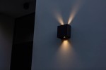 Regulowany kinkiet elewacyjny Gemini Beams w kolorze czarnym, kwadratowy kształt, zamontowany na ścianie. Emituje światło w dwóch kierunkach, tworząc efekt podświetlenia. Tło w neutralnym odcieniu, minimalistyczne otoczenie.
