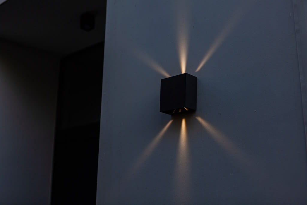 Regulowany kinkiet elewacyjny Gemini Beams w kolorze czarnym, zamontowany na ścianie. Emituje światło w kształcie promieni, tworząc efektowne oświetlenie. Kształt kwadratowy, minimalistyczny design. Tło w odcieniach szarości.