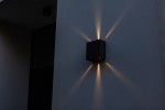 Regulowany kinkiet elewacyjny Gemini Beams w kolorze czarnym, zamontowany na ścianie. Emituje światło w kształcie promieni, tworząc efektowne oświetlenie. Kształt kwadratowy, minimalistyczny design. Tło w odcieniach szarości.