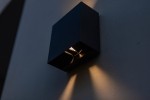 Regulowany kinkiet elewacyjny Gemini Beams w kolorze czarnym, w kształcie sześcianu, zamontowany na ścianie. Emituje światło w dwóch kierunkach, podkreślając teksturę ściany. Widoczne wnętrze lampy, które jest osłonięte z przodu.