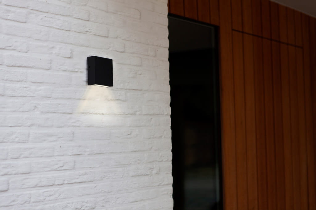 Kinkiet elewacyjny Gemini XF LED w kolorze czarnym, zamontowany na białej, teksturowanej ścianie. Oświetlenie emituje światło w dół, podkreślając minimalistyczny design i prostokątny kształt lampy. W tle widoczna drewniana powierzchnia.