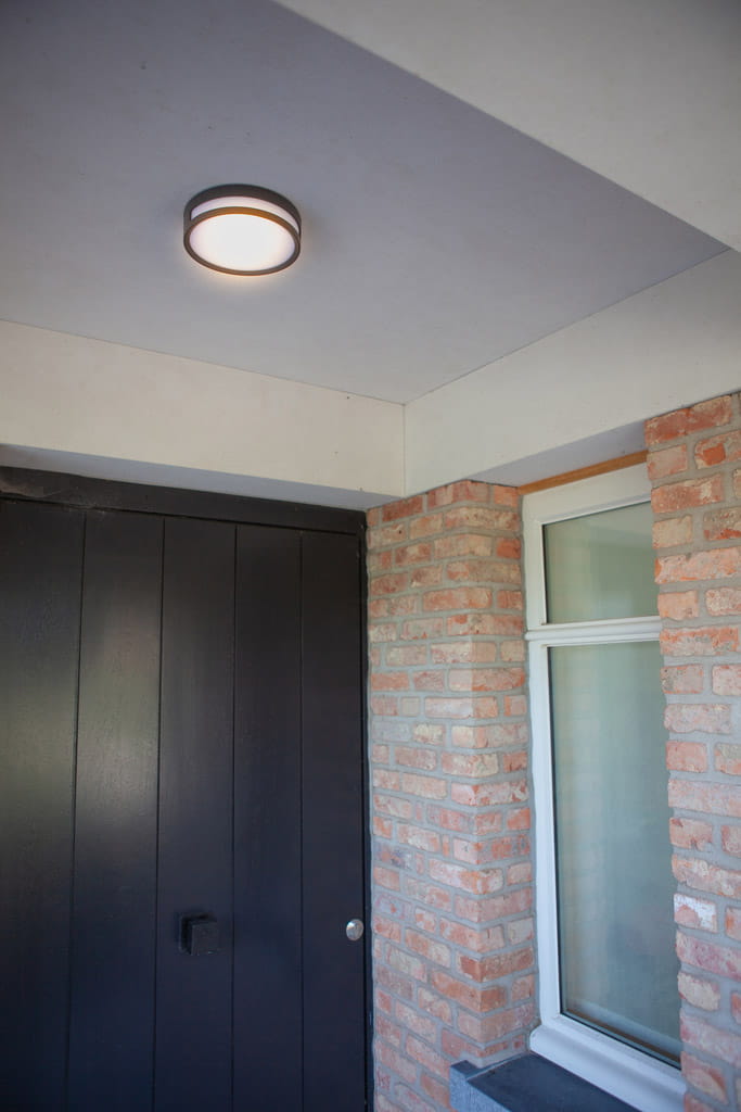 Plafon elewacyjny Rola LED w kolorze czarnym, okrągły z białym kloszem, zamontowany sufitowo. Widoczny w kontekście przy drzwiach, otoczony ceglaną ścianą i oknem, oświetlający wejście.