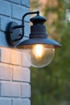 Czarny kinkiet ogrodowy Figo z 1 punktem świetlnym, zamontowany na białej ceglanej ścianie. Lampa z metalowym kloszem i przezroczystą kulą, emitującą ciepłe światło, podkreślająca industrialny styl.