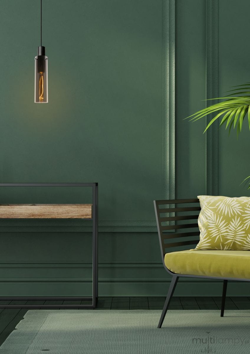 Czarna lampa wisząca Orlando z przezroczystym kloszem, zawieszona na czarnym przewodzie. Widoczna żarówka w środku, otoczona zieloną ścianą. W tle mebel z drewnianym blatem i metalowym stelażem oraz zielona sofa z poduszką w liście.