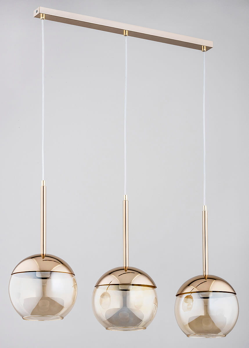 Lampa wisząca 3-punktowa Volda w kolorze złotym. Składa się z trzech okrągłych kloszy w przezroczystym szkłach, zawieszonych na cienkich, białych linkach. Klosze mają metalowe, złote elementy u góry. Montaż sufitowy na prostokątnej listwie.