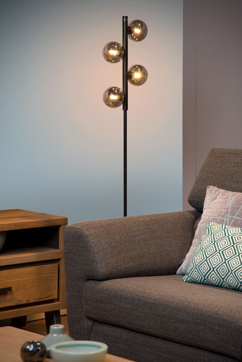 Czarna lampa podłogowa Tycho z czterema szklanymi kulami, umieszczona w pionie. Widoczna obok szarego narożnika i drewnianej szafki. Tło w odcieniach niebieskiego, lampa włącza ciepłe światło, tworząc przytulną atmosferę.