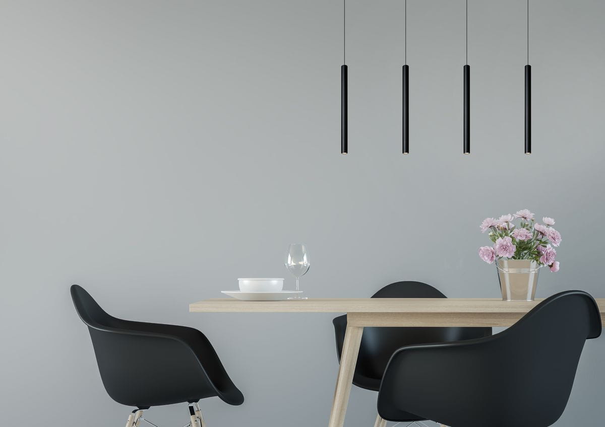 Czarna lampa wisząca LED Lorenz 4-punktowa, składająca się z czterech smukłych, cylindrycznych kloszy, zawieszona nad drewnianym stołem w minimalistycznym wnętrzu. Tło w jasnoszarym kolorze, z kwiatami w wazonie i naczyniami na stole.