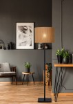 Czarna lampa podłogowa Magius 1-punktowa z okrągłym kloszem z naturalnych włókien, stojąca na prostokątnej podstawie. Ustawiona w nowoczesnym wnętrzu z drewnianą podłogą, roślinami doniczkowymi i obrazem na ścianie.