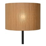 Lampa podłogowa Magius o czarnym stelażu, z okrągłym kloszem w naturalnym kolorze, wykonanym z cienkich pasków materiału. Ujęcie frontalne, lampa stoi na podłodze, emitując ciepłe światło przez klosz.