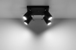 Czarna lampa sufitowa (spot) 4-punktowa, z czterema cylindrycznymi reflektorami skierowanymi w różne strony. Ujęcie z góry, na jednolitym tle. Reflektory mają jasne źródło światła, montowane do prostokątnej podstawy.