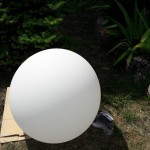 Biała lampa ogrodowa Cumulus L, kształt kulisty, umieszczona na kartonie na trawie, z widocznymi elementami mocującymi. Tło z roślinnością, lampa przygotowana do ustawienia w ogrodzie.