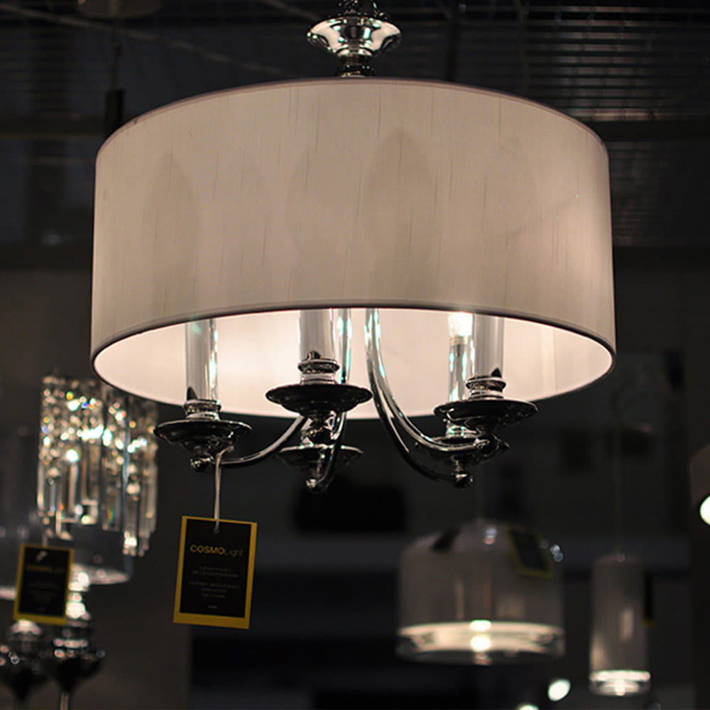 Lampa wisząca Abu Dhabi 5-punktowa w kolorze chromu z białym, okrągłym abażurem. Posiada pięć źródeł światła, z metalowymi uchwytami. Widoczna etykieta z nazwą producenta. Produkt zawieszony w przestrzeni, w tle inne lampy.