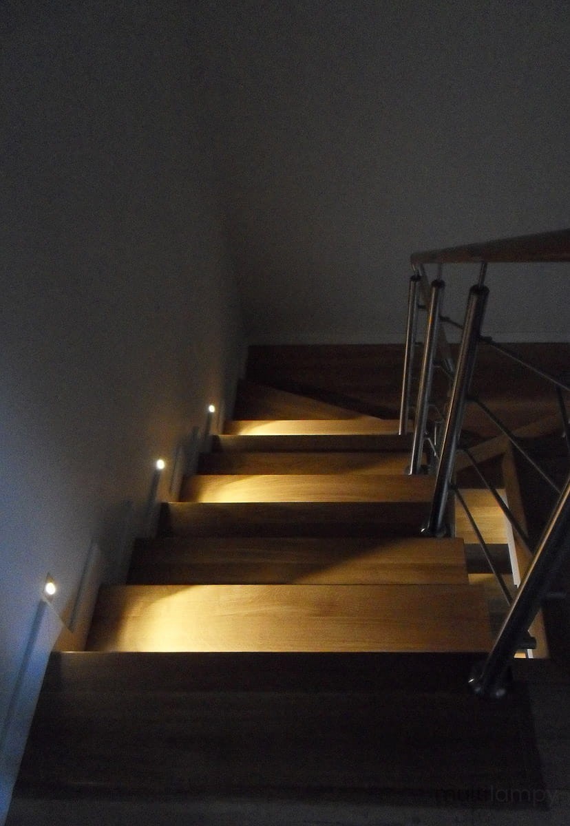 Lesel LED oprawa schodowa 001 L, umieszczona na schodach, emitująca ciepłe światło, wkomponowana w drewniane stopnie. Widoczna metalowa balustrada po prawej stronie, ciemne tło dodaje kontrastu.