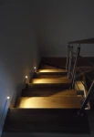 Lesel LED oprawa schodowa 001 L, umieszczona na schodach, emitująca ciepłe światło, wkomponowana w drewniane stopnie. Widoczna metalowa balustrada po prawej stronie, ciemne tło dodaje kontrastu.