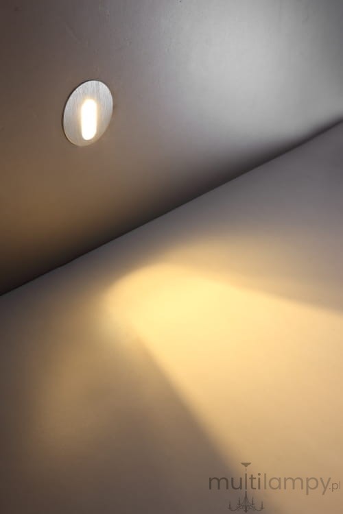 Lesel LED oprawa schodowa 002, okrągła, umieszczona w ścianie. Emituje ciepłe światło, tworząc delikatny cień na podłożu. Prosta, minimalistyczna konstrukcja w kolorze srebrnym, idealna do oświetlenia schodów.