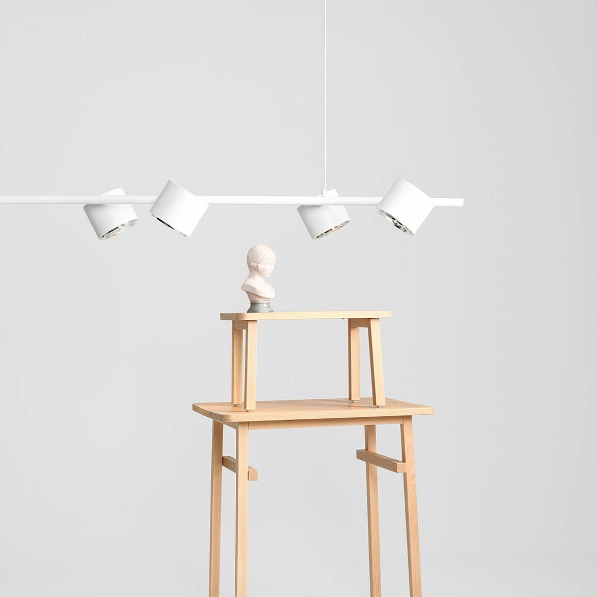Lampa wisząca Bot 6-punktowa w białym kolorze, zawieszona na cienkim przewodzie, z cylindrycznymi kloszami w różnych kierunkach. Na drewnianym stoliku znajduje się rzeźba popiersia. Tło proste, jasne.