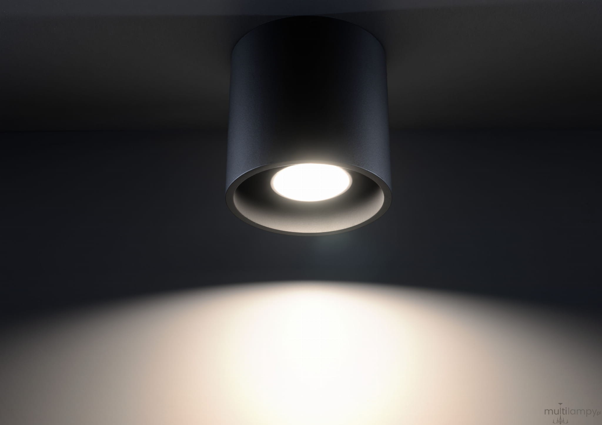 Czarna lampa sufitowa Orbis 1-punktowa, o cylindrycznym kształcie, zamontowana na suficie. Źródło światła umieszczone w centrum, emitujące jasne światło, które tworzy delikatny cień na ścianie. Proste, minimalistyczne wzornictwo.