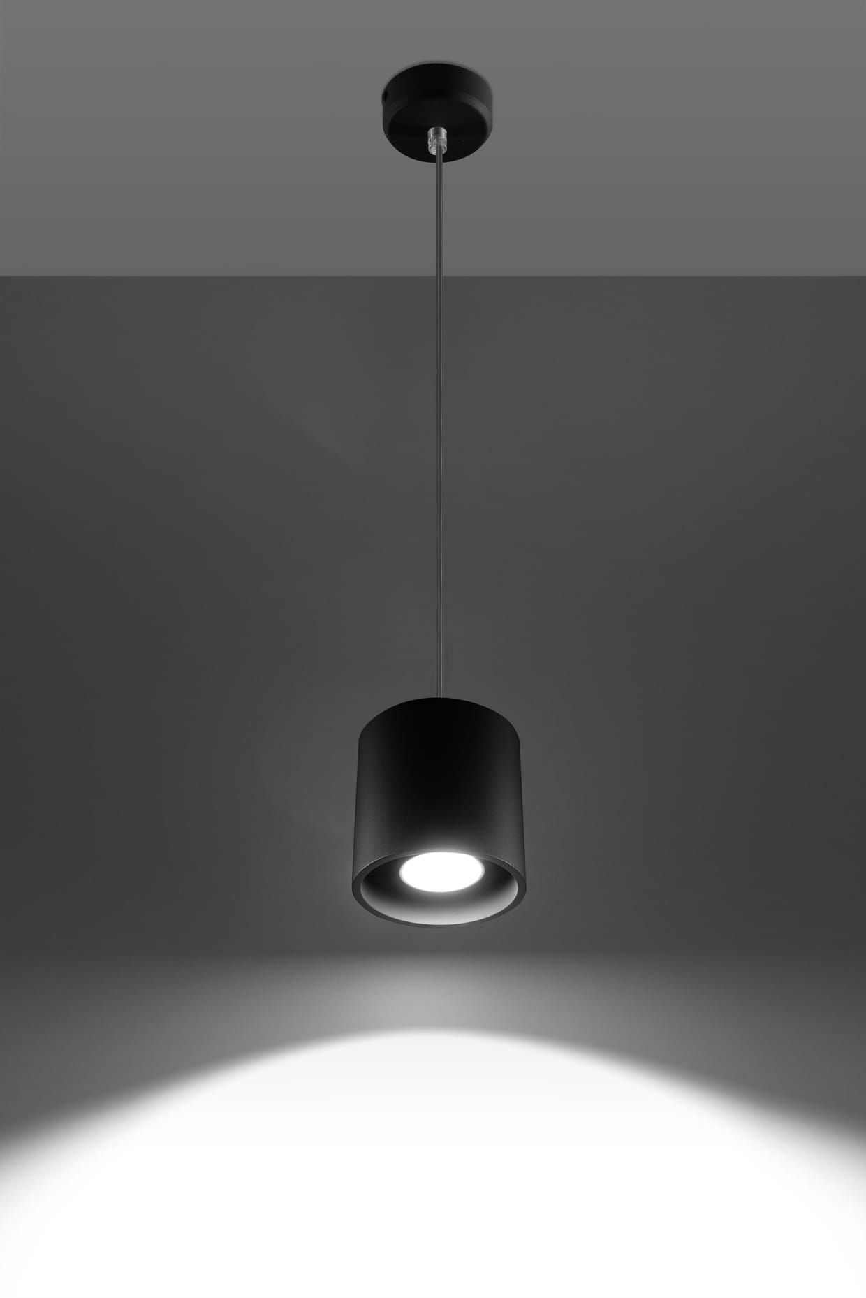 Czarna lampa wisząca Orbis SL.0051, w kształcie cylindrycznym, zawieszona na cienkim przewodzie. Ujęcie z dołu, pokazujące źródło światła oraz wyraźny cień na ścianie. Prosty design, idealna do nowoczesnych wnętrz.