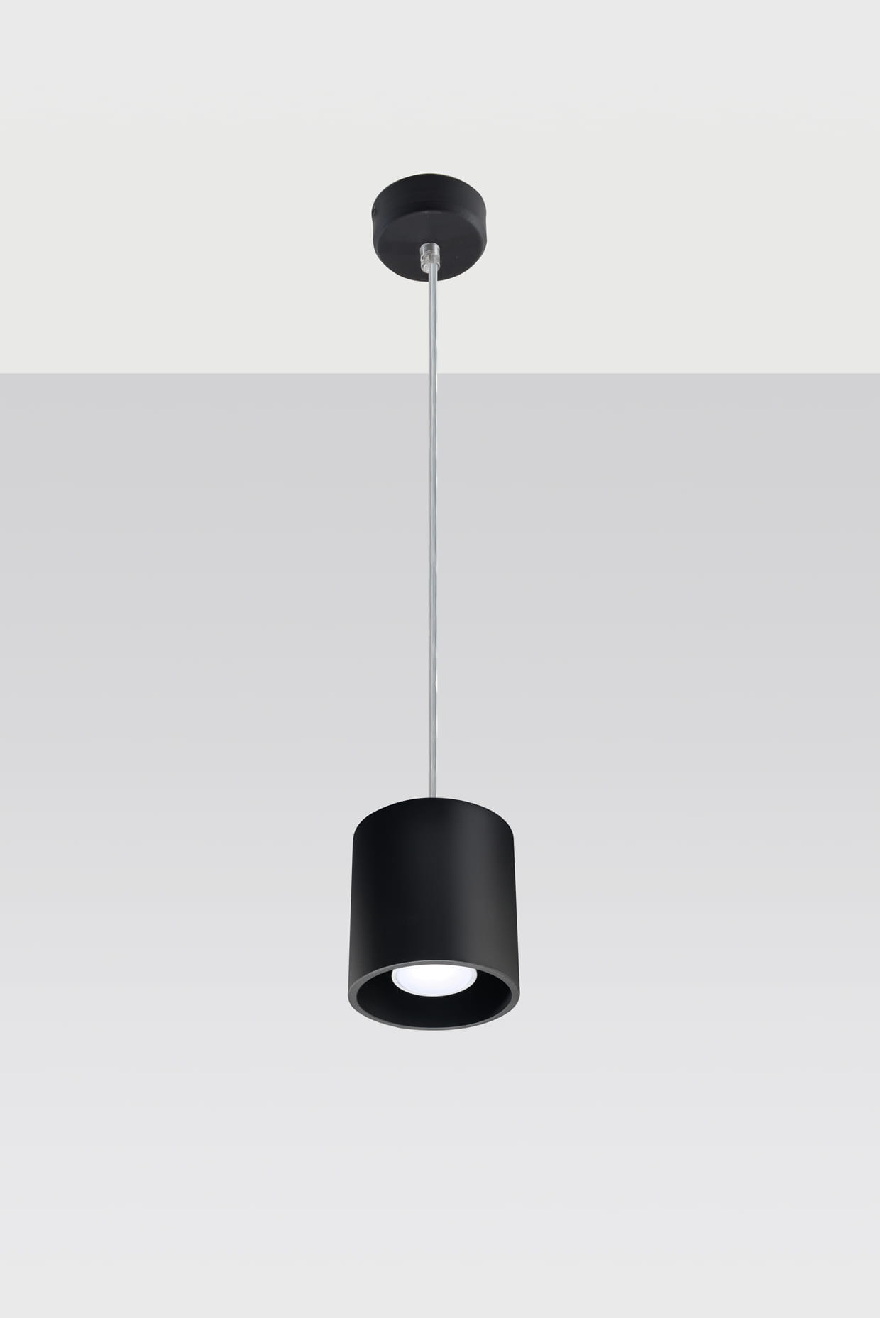 Czarna lampa wisząca Orbis, 1-punktowa, o cylindrycznym kształcie. Zawieszona na cienkim, metalowym przewodzie. Prosta, matowa powierzchnia z okrągłym otworem na źródło światła. Tło jasne, jednolite.