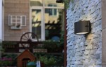 Kinkiet elewacyjny Bonn w kolorze ciemnoszarym, montowany na ścianie, o cylindrycznym kształcie. Oświetlenie skierowane w dół, zamontowane na tle kamiennej elewacji, w otoczeniu roślinności i drewnianych elementów architektury.