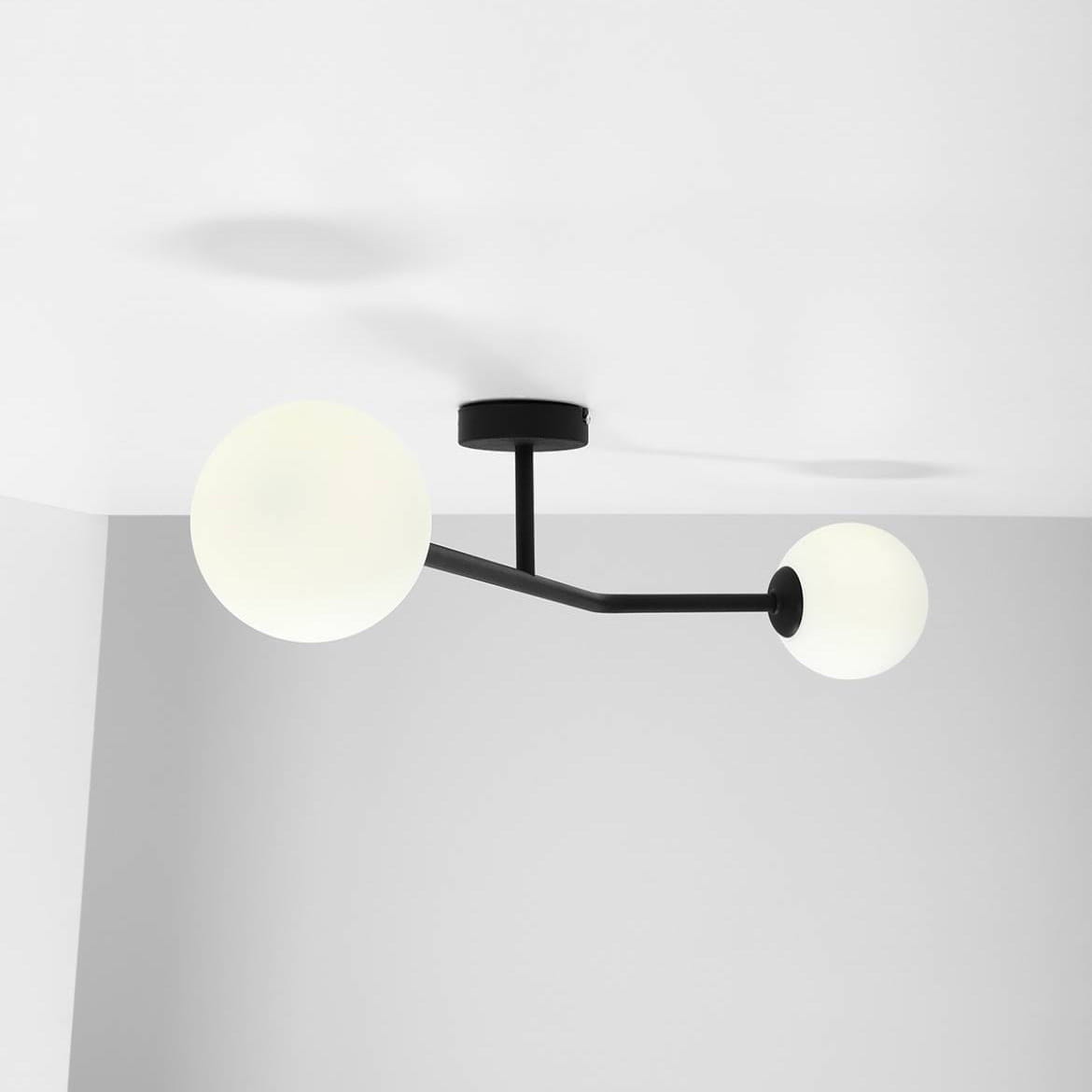 Lampa sufitowa 2-punktowa w kolorze czarnym. Konstrukcja z matowego metalu z dwoma okrągłymi, białymi kloszami. Ujęcie z dołu, ukazujące zamontowaną lampę na białym suficie, z jednym ramieniem skierowanym w lewo, a drugim w prawo.